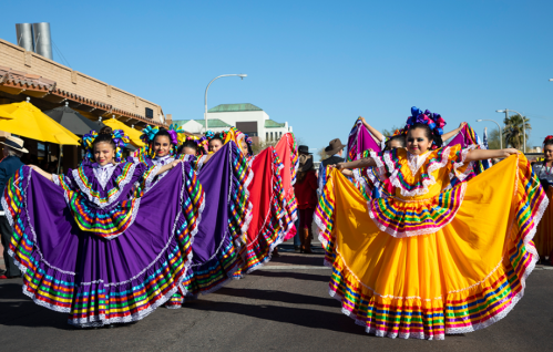 A vibrant parade featuring dancers in colorful traditional dresses, showcasing cultural celebration under a clear blue sky.