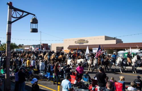 A crowd watches a parade with horses and riders in front of a market building under a clear blue sky.