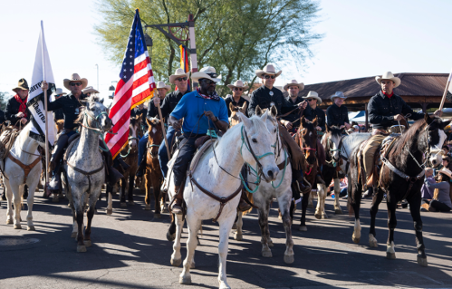 A group of cowboys on horseback, led by a man holding an American flag, parades down a street.