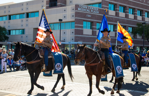 Three mounted riders in uniform carry flags during a parade in front of a medical plaza. Crowds watch from the sidelines.