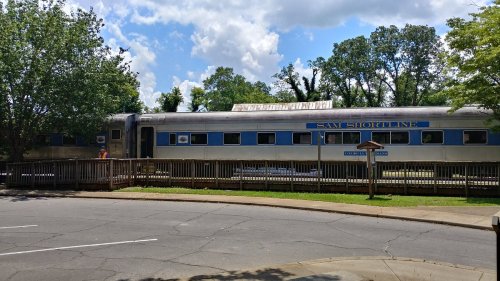 A vintage train car named "Sam Shortline" parked beside a wooden platform, surrounded by trees and blue skies.