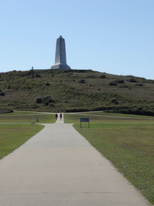 A paved path leads to a tall monument atop a grassy hill under a clear blue sky. Two people walk towards it.