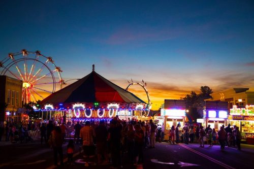 A vibrant carnival scene at dusk, featuring a Ferris wheel, colorful lights, and a crowd enjoying the festivities.