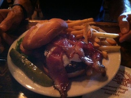 A close-up of a burger topped with bacon and sauce, served with fries and a pickle on the side.