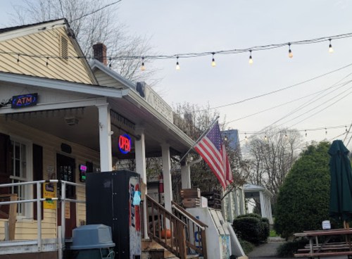 A cozy building with an open sign, an American flag, and string lights, surrounded by greenery and outdoor seating.