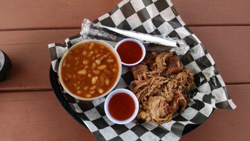 A plate with pulled pork, baked beans, and two cups of sauce, served on checkered paper.