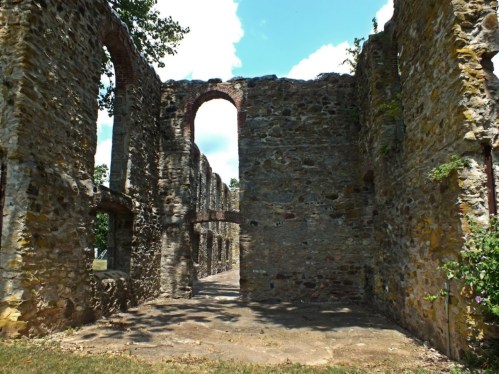 Ruins of a stone building with arched openings, surrounded by greenery and under a bright blue sky.