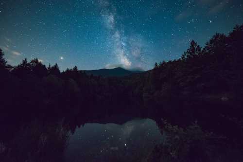 A serene night scene featuring a starry sky and the Milky Way reflected in a calm lake surrounded by trees.