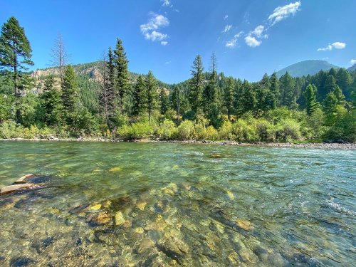 A clear river flows through a lush green landscape, surrounded by tall trees and mountains under a blue sky.