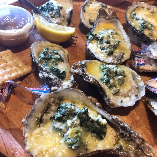 A wooden platter with baked oysters topped with green herbs, a lemon wedge, crackers, and a small cup of sauce.