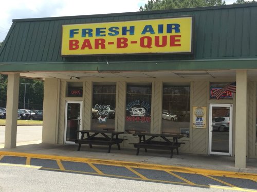 Exterior of a barbecue restaurant with a bright sign, picnic tables outside, and an open sign in the window.