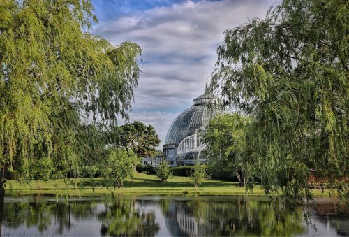 A glass conservatory surrounded by lush greenery and a calm pond, under a partly cloudy sky.