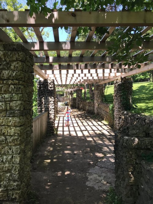 A stone pathway with a wooden trellis overhead, casting shadows, surrounded by greenery. A person walks in the distance.