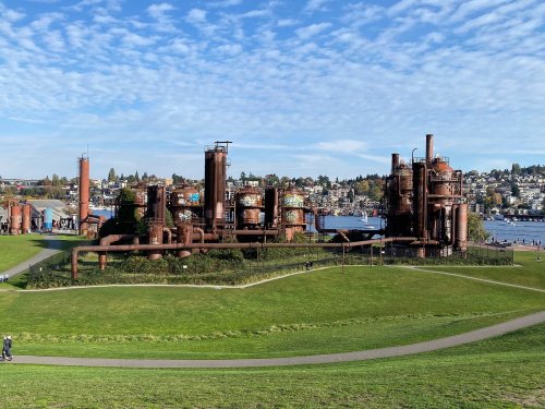 Industrial structures with rusted pipes and tanks, set against a backdrop of green grass and a blue sky.