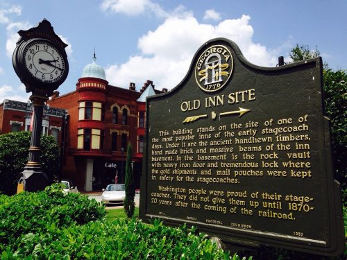 Historic sign for the Old Inn Site in Georgia, with a clock and red brick buildings in the background.