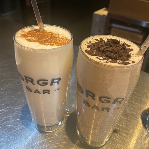 Two tall glasses of milkshakes, one topped with cinnamon and the other with crushed cookies, on a metal table.