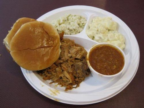 A plate with two sandwich buns, pulled pork, rice, potato salad, and a cup of chili.