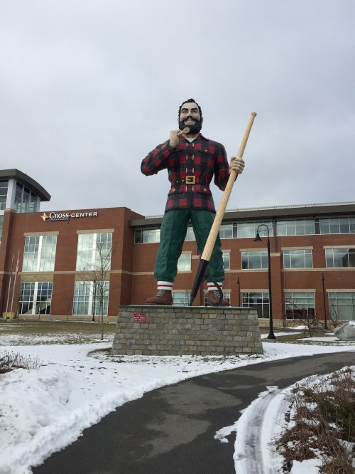 A large statue of a lumberjack in plaid stands on a stone pedestal outside a modern building, with snow on the ground.