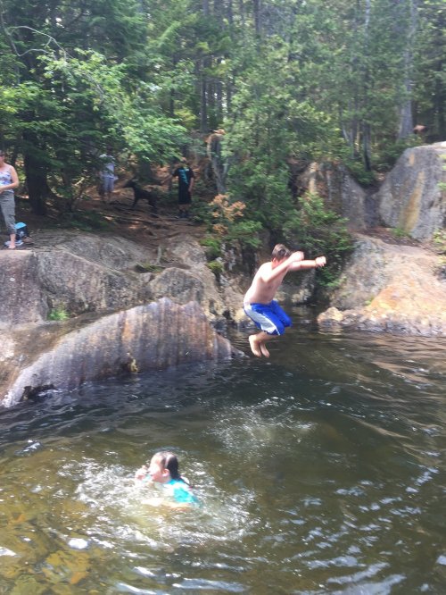A person jumps into a lake while another swims nearby, surrounded by trees and rocky terrain.