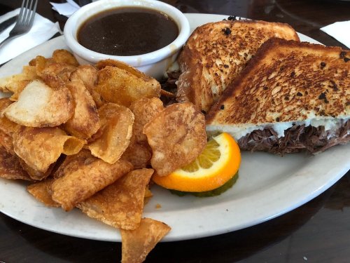 A plate featuring a toasted sandwich with beef, a side of crispy potato chips, and a small bowl of dark gravy.