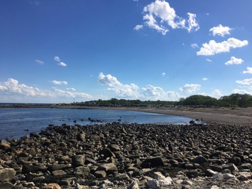 A rocky shoreline under a blue sky with scattered clouds, leading to calm waters and distant greenery.