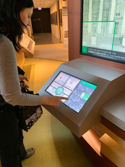 A woman interacts with a touchscreen display in a museum, exploring information about paper money.