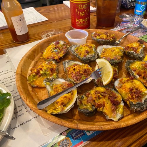 A wooden platter filled with baked oysters topped with cheese, bacon, and a lemon wedge, served with a dipping sauce.