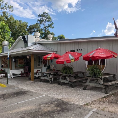 A quaint restaurant with outdoor seating under red umbrellas, surrounded by trees and a clear blue sky.