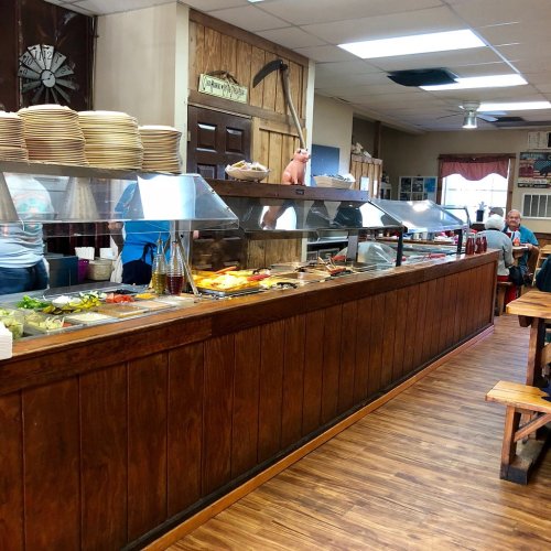 A rustic buffet with a wooden counter, food trays, and diners seated at wooden tables in a casual restaurant setting.