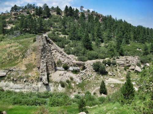 Ruins of a stone structure on a hillside, surrounded by lush greenery and trees under a blue sky.