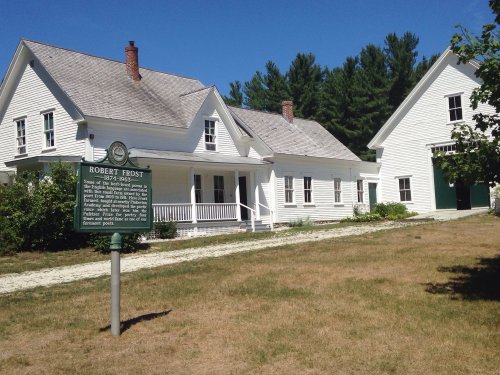 A white house with a green historical marker in front, surrounded by grass and trees on a sunny day.