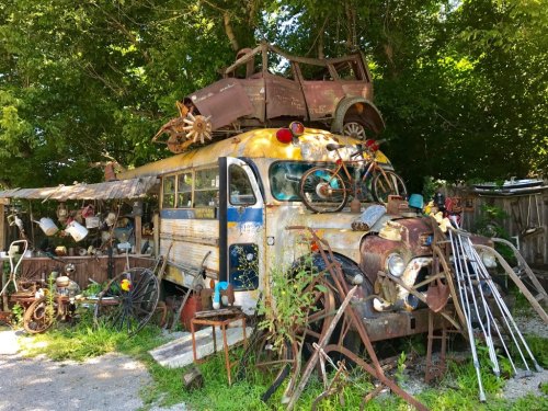 A rusty, vintage bus topped with an old car, surrounded by various discarded items and greenery.