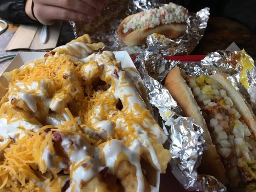 A close-up of loaded fries topped with cheese and sour cream, alongside a hot dog in foil with toppings.