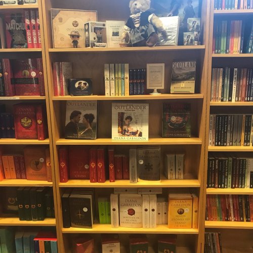 A wooden bookshelf filled with colorful books, including several titles by Diana Gabaldon, and a teddy bear on top.