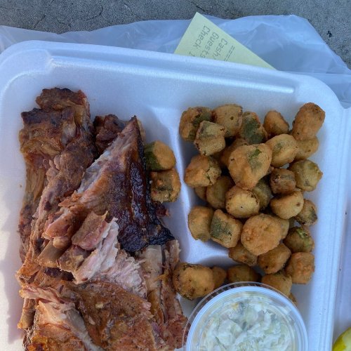 A takeout container with sliced barbecue meat, fried okra, and a small cup of coleslaw on a textured surface.