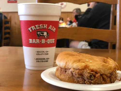 A BBQ sandwich on a plate next to a large drink cup, with a casual dining setting in the background.