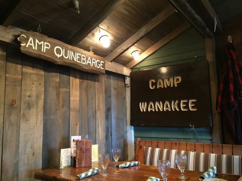 Interior of a rustic cabin with wooden walls, featuring signs for "Camp Quinebarge" and "Camp Wanakee."