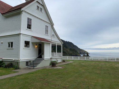 A white house with a red roof overlooks a calm ocean, surrounded by green grass and a scenic coastal landscape.