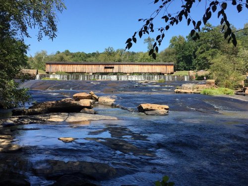 A serene landscape featuring a wooden structure over a waterfall, surrounded by trees and rocky terrain under a clear blue sky.