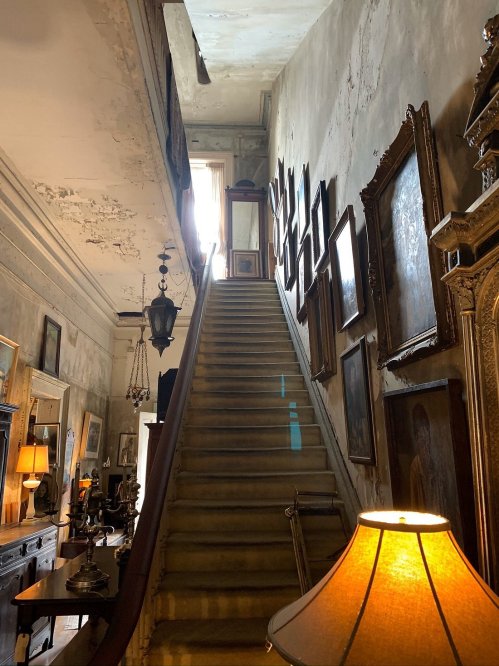 A dimly lit staircase leads upward, flanked by vintage paintings and a warm lamp, in a weathered, historic interior.