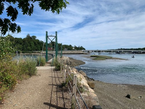A scenic view of a bridge over a river, with a pathway lined by greenery and boats in the distance under a cloudy sky.