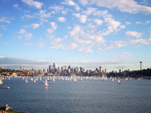 A scenic view of a city skyline with sailboats on the water under a blue sky with scattered clouds.