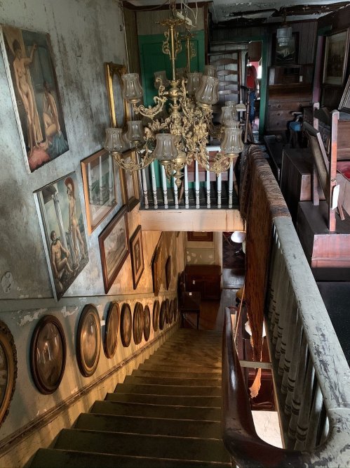A vintage staircase with ornate chandelier, lined with framed artwork and mirrors, leading to an upper level.