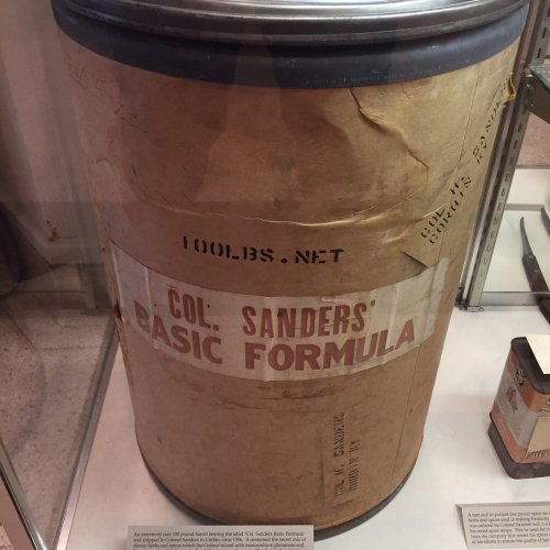 A large, vintage container labeled "COL. SANDERS' BASIC FORMULA" displayed in a museum.