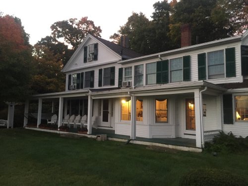 A large white house with green shutters, featuring a porch and illuminated windows, surrounded by trees at dusk.