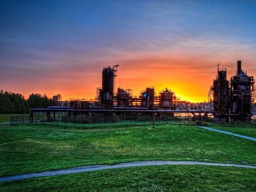 Sunset over an industrial landscape with silhouettes of structures against a colorful sky and green grass in the foreground.