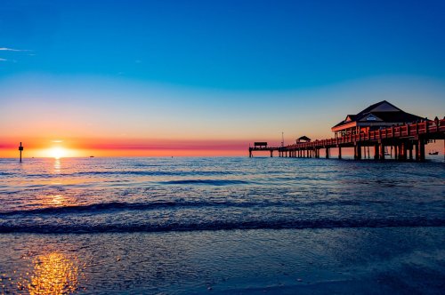 A serene sunset over the ocean, with a pier silhouetted against vibrant orange and blue skies.