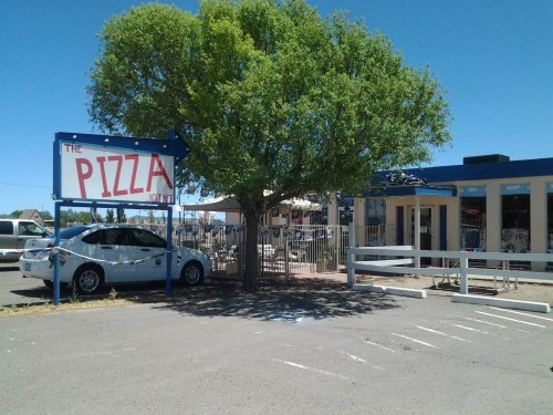 A pizza restaurant with a large sign, outdoor seating, and a tree in front, set against a clear blue sky.