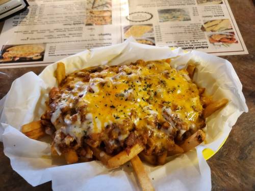 A plate of loaded fries topped with melted cheese, chili, and herbs, served in a white paper basket.