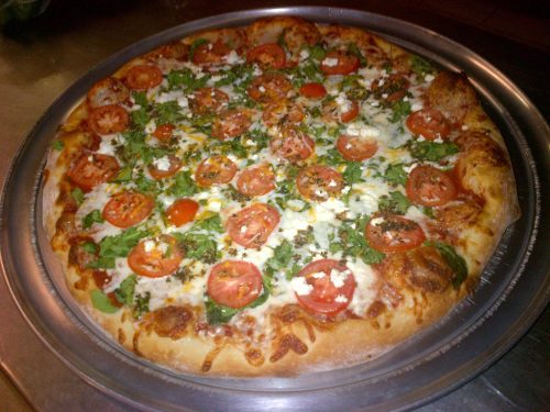 A freshly baked pizza topped with sliced tomatoes, greens, and cheese on a golden crust, served on a metal tray.